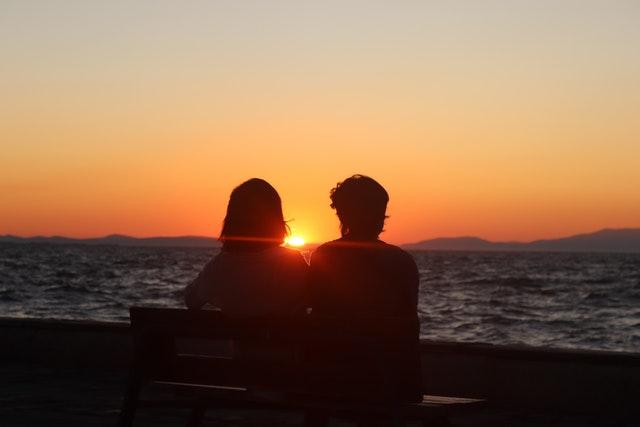 Un couple est assis face à la mer sur un banc, entre eux le soleil se couche à l'horizon.