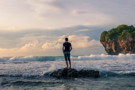 Un homme se tient debout sur un rocher en bord de mer, il est cerné par les vagues et regarde la mer.