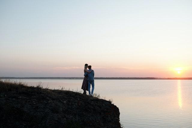 Un couple est enlacé en haut d'une falaise, ils regardent la mer et le soleil qui se couche.