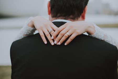 un homme en costume noir de dos, sur son cou les mains d'une femme habillée en mariée.