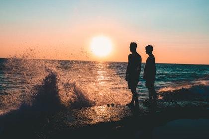 Un soleil couchant au bord de la mer, la silhouette d'un couple se dessine devant une vague