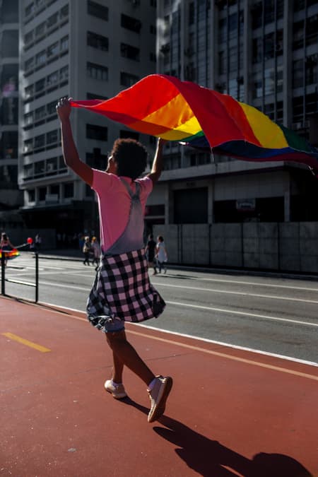 Une personne marche sur un trottoir en ville en tenant au dessus de sa tête un drapeau arc-en-ciel.