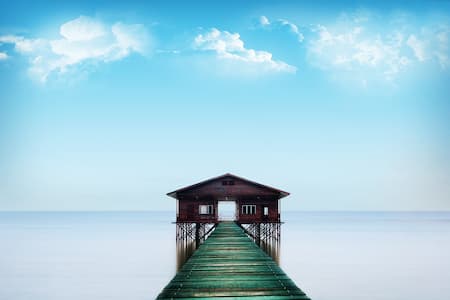 Un panorama de mer et ciel bleu, en face se trouve un ponton qui mene à une maison de bois sur pilotis