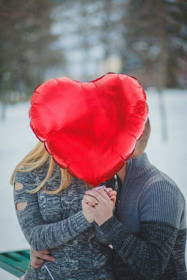 Un couple s'embrasse en tenant devant leurs visages un gros ballon rouge en forme de coeur.