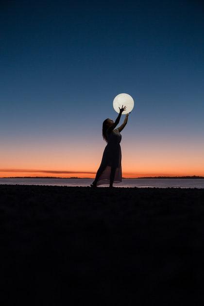 Une femme au crépuscule lève les mains et donne l'impression d'attraper la pleine lune.